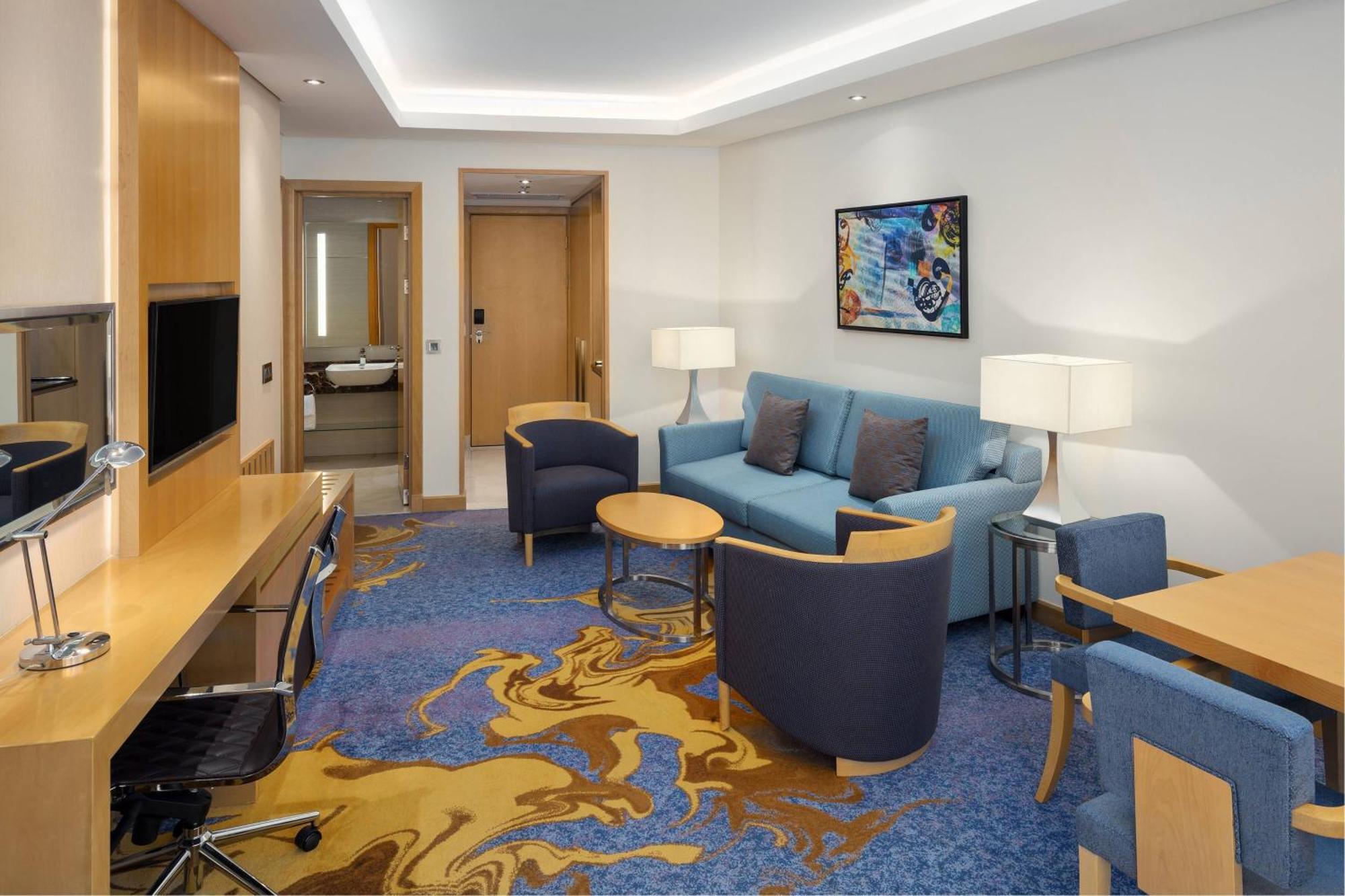 Sheraton Makkah Jabal Al Kaaba Hotel Mekka Kültér fotó The photo displays a modern hotel room or suite. The room features a comfortable seating area with a blue couch and two blue armchairs surrounding a small round table. A wooden desk with a black office chair is positioned against one wall, accompanie