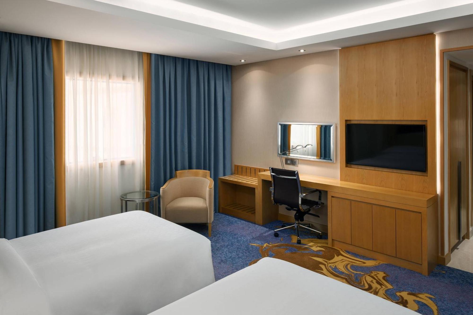 Sheraton Makkah Jabal Al Kaaba Hotel Mekka Kültér fotó The photo shows a hotel room featuring two beds with white linens. There are large windows covered with blue curtains that allow natural light to enter. On one side of the room, there is a wooden workstation with a chair and a mirror, along with a fl