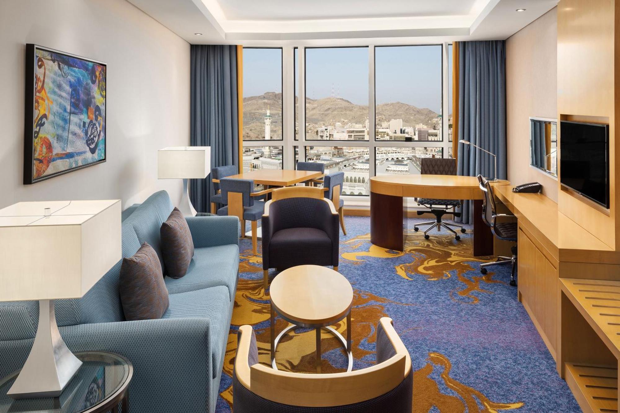Sheraton Makkah Jabal Al Kaaba Hotel Mekka Kültér fotó The photo shows a well-decorated hotel room featuring a modern and comfortable interior. There is a cozy seating area with a blue sofa and armchairs, complemented by a round coffee table. A desk with a chair is also present, ideal for work. The room 