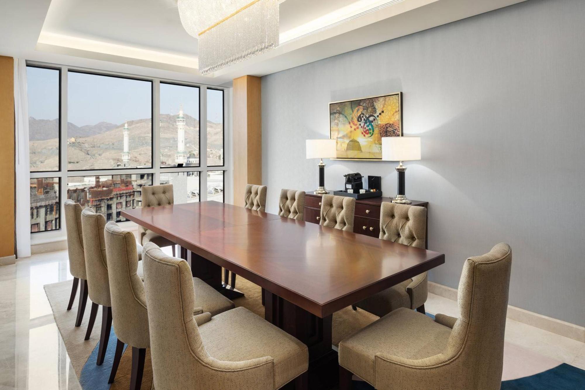 Sheraton Makkah Jabal Al Kaaba Hotel Mekka Kültér fotó The photo shows a modern conference room with a large wooden table surrounded by upholstered chairs. The room is well-lit, featuring large windows that offer a view of the outdoors, possibly with mountains in the background. On one wall, there is a p