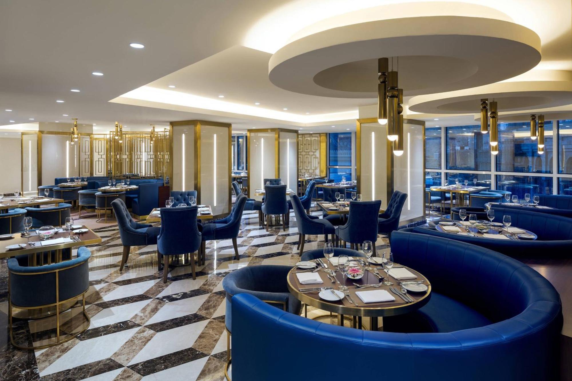Sheraton Makkah Jabal Al Kaaba Hotel Mekka Kültér fotó The photo shows the interior of a modern restaurant. The design features elegant curved blue seating arranged around round tables with plates and glassware set for dining. The flooring is a striking marble pattern, adding to the upscale ambiance. The